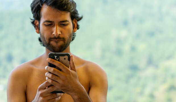 Selective focus chest up shot of a shirtless young Indian handsome man looking mischievously at his smartphone.