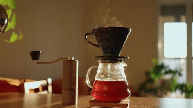 Morning sunlight shining on a home kitchen table where freshly brewed filter coffee is dripping into a glass server, with steam rising from the dripper and a manual grinder standing nearby