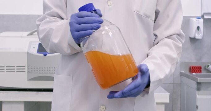 A full-length view of a chemist holding a large chemical bottle containing an orange substance and swirling the reagent inside the container. Mixing of chemical substances.