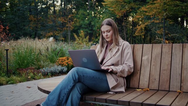 Blonde woman works on laptop while sitting on wooden park bench