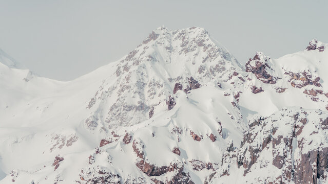 Snow-covered rocky mountain slopes with exposed dark rock under pale overcast sky