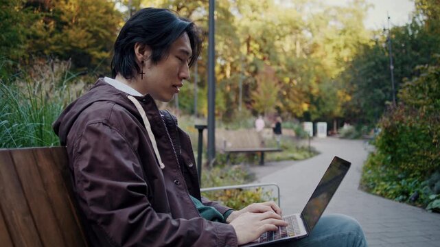 Focused Asian man types on laptop during outdoor park work