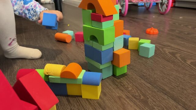 A child reaches for a yellow block beside a colorful tower on a wooden floor, construction play, childhood learning, motor skills.