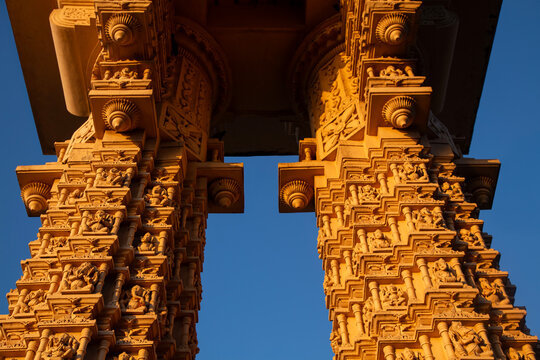 Intricately Carved Stone Temple Pillars with Ganesha Idols at Golden Hour. Vertical View of Ornate Hindu Temple Gateway Columns Against Blue Sky and Palm Trees