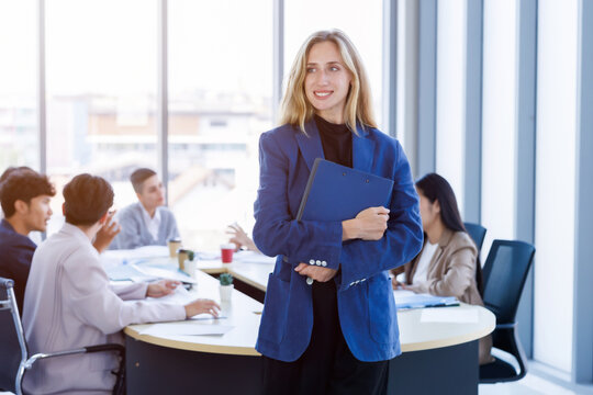 Confident Caucasian businesswoman holding document folder wearing a blue blazer and holding a blue folder while smiling at the camera team of colleagues working around a meeting in corporate office.