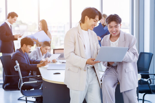 Two young Asian businessmen discussing work on a laptop computer Background features a diverse team of colleagues working and discussing documents in a bright, modern corporate office environment.