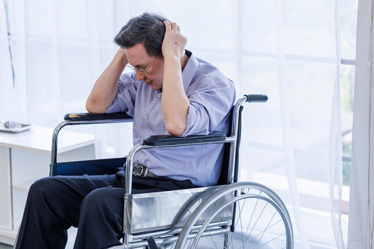Stressed elderly Asian man sitting in wheelchair holding his head in despair indoors.representing mental health issues, elderly depression, loneliness, or chronic illness in seniors.