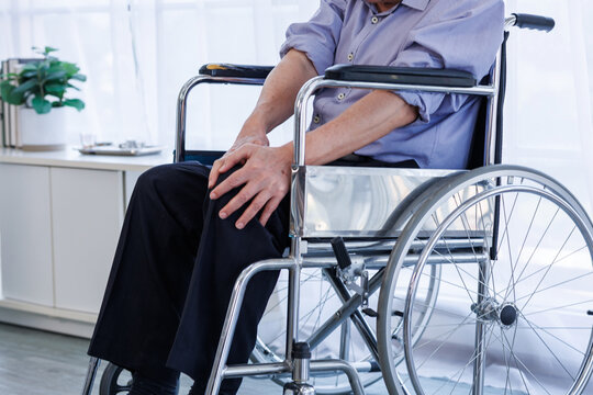 Close-up shot focusing on an elderly man sitting in a wheelchair, holding his aching knee with both hands due to joint pain, arthritis, or injury. in a home care or hospital environment.