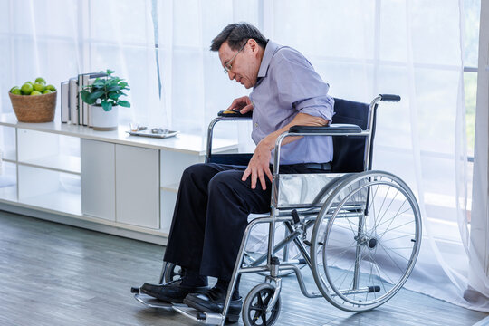 A middle-aged Asian man, a patient sitting in a wheelchair, holding his aching knee with both hands due to joint pain, arthritis, or injury. in a home care or hospital environment.