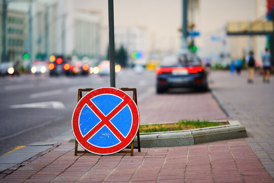 Circular no stopping and no parking traffic sign standing on paved city sidewalk to regulate urban vehicle traffic with blurred street background. Temporary prohibition road sign on pedestrian walkway