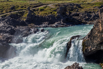 Turquoise rapids rush over dark rock ledges in the Skjalfandafljot River below Godafoss in North Iceland, with foaming cascades and drifting spray beneath grassy banks. © Artem