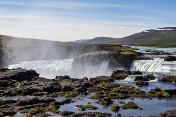 Powerful cascades at Godafoss send mist above dark rocks as the Skjalfandafljot River surges through North Iceland, with mossy pools and snow-streaked hills under a blue summer sky. © Artem