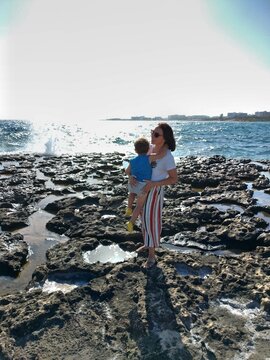 A woman holds a child on a dark rocky shoreline against sparkling water, combining a family holiday portrait with bright coastal light