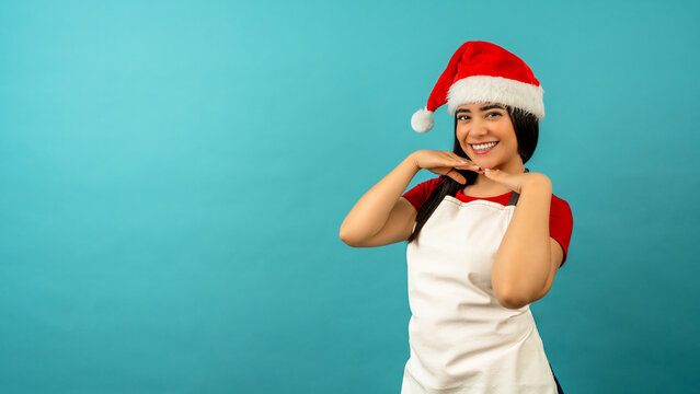 Woman in a santa hat and white apron smiling and posing against a bright turquoise background