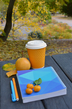 Coffee to go in orange paper cup with lid next to ripe pear ,blue notebook , pens arranged on dark wooden table against background of autumn park. Morning or afternoon cup of baverage in park cafe .