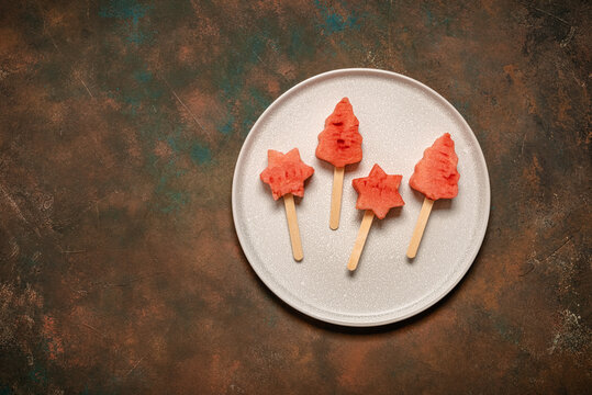 Watermelon pieces in the shape of Christmas tree and star on a stick, watermelon popsicles, summer dessert, dark rustic background, top view, flat lay, copy space.
