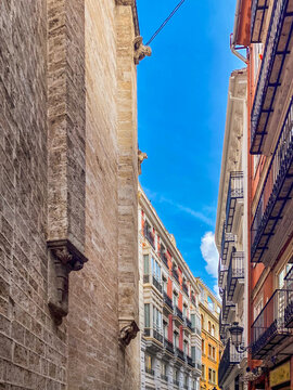Narrow pedestrian street in the old town of Valencia, Spain with the ancient stone wall of Gothic church contrasting with colorful residential apartments  with iron balconies under blue summer sky