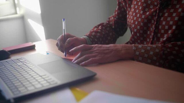 Woman's hands writing notes next to a laptop at an office desk, close-up, slow motion