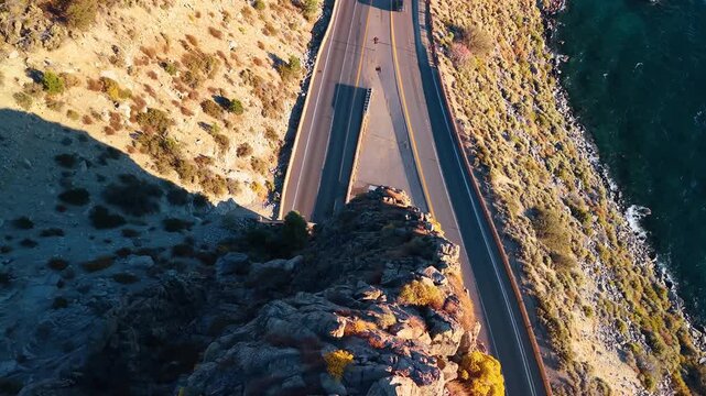 A lakeside road passes behind a jagged rock outcrop. A broad shadow crosses the asphalt beside sunlit stone and dark blue water in an overhead view.