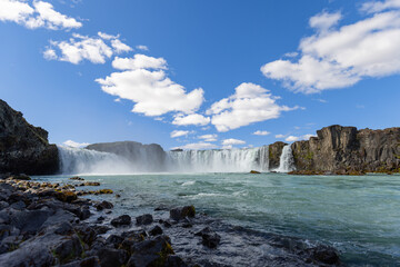 Seen from the rocky river edge, Godafoss spreads across the frame as white cascades meet churning turquoise water beneath scattered clouds in the summer sky of North Iceland. © Artem