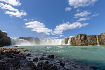 From a low, rocky bank, Godafoss spans the view with broad white cascades and rising mist above the turquoise Skjalfandafljot River on a sunny summer day in North Iceland. © Artem