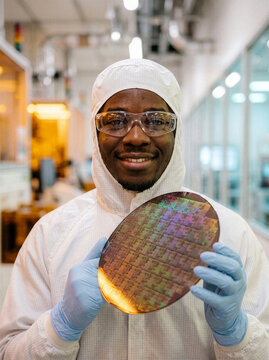 African American semiconductor technician holding processed silicon wafer in cleanroom for microchip manufacturing.