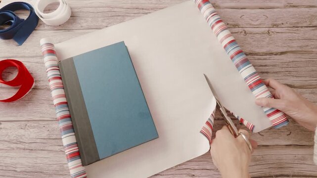 Hands cutting striped wrapping paper for wrapping a book, and white, blue and red ribbons on wooden table. Real time video.
