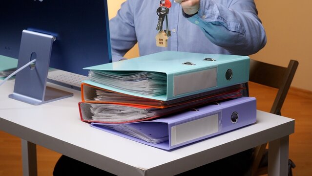 Office professional holding house keys above stacked paperwork, organizing real estate documents at a desk with computer, suggesting home ownership, mortgage processing and property finance