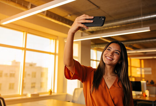Smiling young woman taking selfie with smartphone in modern office during golden hour with warm natural sunset light