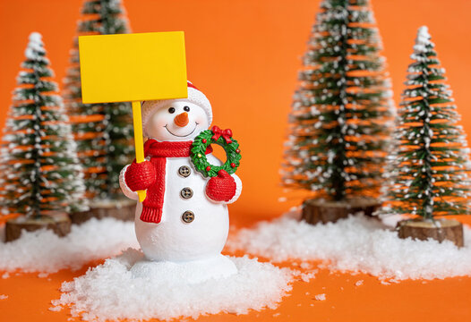 Snowman figurine holding blank yellow sign and wreath on artificial snow with orange background and miniature pine tree