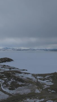 High above Baikal, an expansive sheet of winter ice meets a rugged, snow-covered shore beneath heavy clouds, capturing the austere scale and stillness of Russia&rsquo;s remote Siberian wilderness