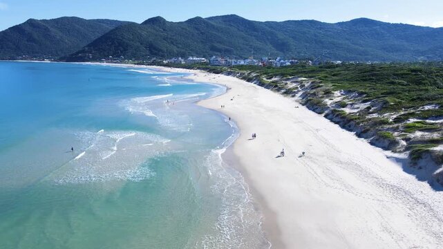 Drone aerial view of Praia dos Acores coastal sand dunes and pristine beach in Florianopolis, Brazil, featuring white sand shoreline, turquoise ocean waves, and green mountains background
