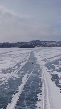 A winter route crosses Lake Baikal's frozen expanse, marked across pale, snow-dusted ice and leading toward distant mountain ridges beneath an overcast sky in Irkutsk Oblast, Russia