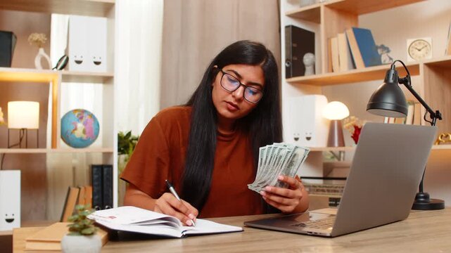 Indian woman on home table plans a big purchase and writes budget notes while comparing costs. Arabian girl finds an affordable option and nods with satisfaction after making a smart financial choice.