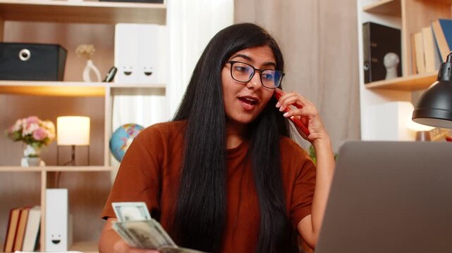 Indian woman on home table calls seller to discuss a large purchase while counting cash and planning budget. Arabian girl asks seller about price before buying costly item online with laptop at home.
