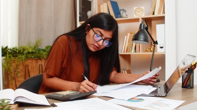 Indian woman on home table highlights wrong data in documents during budget check and checks figures with calculator. Arabian girl looks focused at home while correcting mistake in financial records.