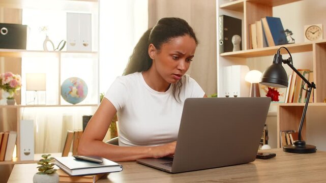 African American young woman on home table browses job vacancies on laptop and reads requirements with focused expression. Black girl compares career options and checks notes while searching for work.