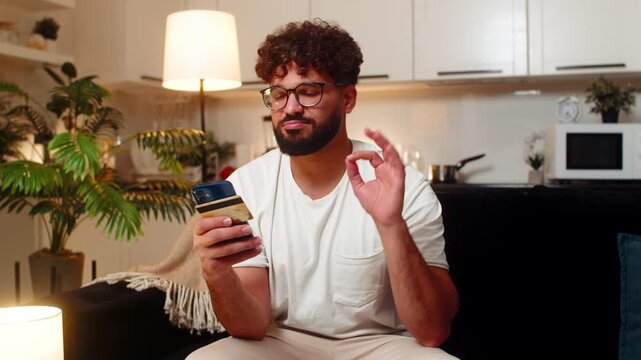 Arabian man at home uses smartphone and card confirms payment checks promo code sees discount makes ok sign. Egyptian guy on sofa saves receipt smiles feels safe secure purchase done at night indoors.