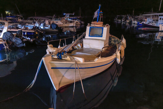 A small traditional Greek fishing boat is moored in dark, tranquil harbor waters at night, surrounded by other boats gently reflecting in the quiet sea