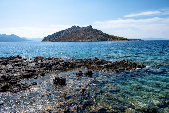 Clear turquoise waters lap against the rugged rocky shore near Perdika village on Aegina Island, offering a scenic view of Moni Island and distant sailboats in the Saronic Gulf.
