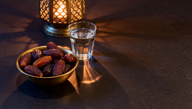 Iftar dates and water still life, medjool dates in a brass bowl with a glass of water and a glowing lantern on a dark table