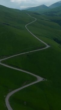 Drone footage follows a twisting paved road across lush Caucasus hills near Aktoprak, Kabardino-Balkaria, Russia, where open grass slopes meet hazy peaks beneath a shifting blanket of clouds