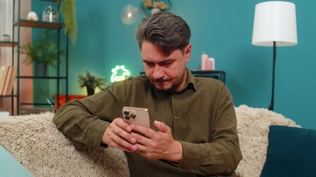 Middle-aged man at home types smartphone message pauses erases rewrites showing hesitation and thought about words. Young adult guy on sofa holds mobile phone pondering decision seeking best wording