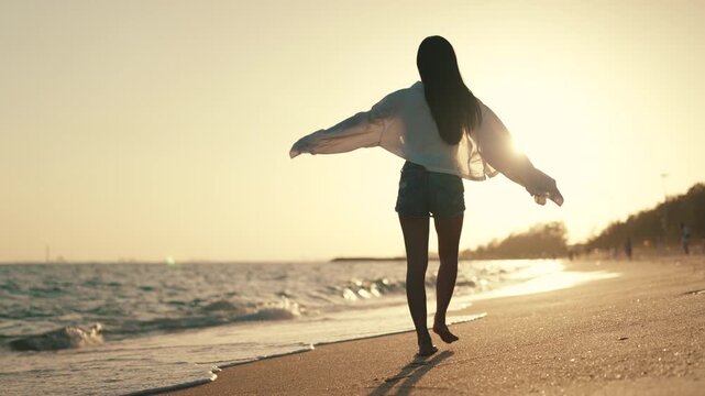 Summer beach freedom, woman walking barefoot along seashore at golden sunset with arms outstretched