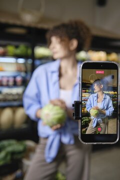 Woman recording video in grocery store using her smartphone. Shelves display various vegetables in background. Focus on sharing knowledge about healthy eating and vegetable benefits