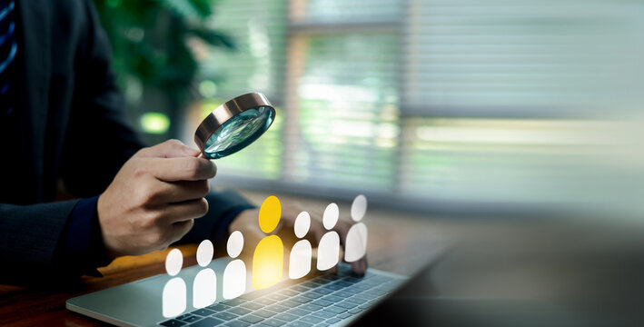 Business person holding a magnifying glass examining a highlighted candidate icon among a team of people over a laptop. Concept of human resources, recruitment, talent acquisition, and hiring.