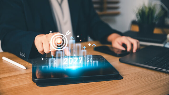 Businessperson touching digital target icon and 2027 growth chart on tablet screen, planning corporate business strategy, forecasting annual financial performance, and achieving future success goals