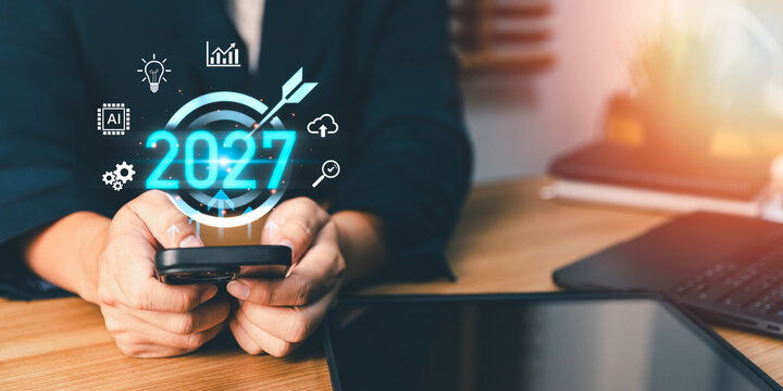 Businessperson using smartphone with 2027 digital target icon and technology symbols, planning future corporate strategy, innovation, and aiming for annual business growth success