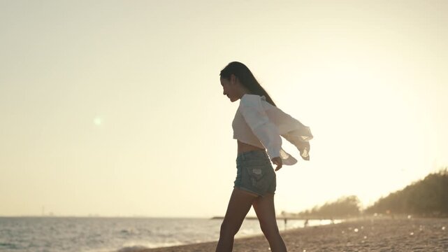 Summer vacation freedom on beach at sunset, Asian woman twirling with arms outstretched and white shirt flowing in the wind by the sea