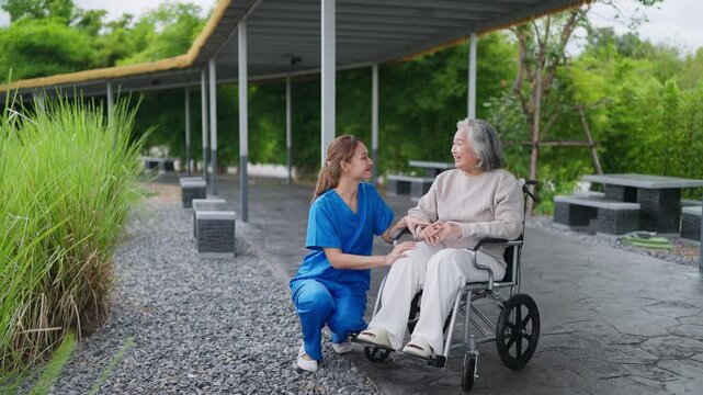 Senior woman smiles gently in a wheelchair. Caregiver kneels beside her, hands clasped warmly. Both share a quiet, comforting moment outdoors